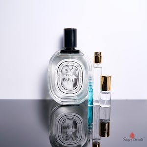 May include: A clear glass perfume bottle with a black cap and a white oval label reading L'EAU PAPIER 34 boulevard saint germain parisse, accompanied by three smaller glass sample vials with gold and blue accents, arranged on a reflective surface.