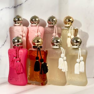 May include: An assortment of perfume bottles in various shades of pink, peach, and amber. Each bottle has a decorative silver or gold cap and a tassel. The bottles are arranged on a white surface with a marble background.