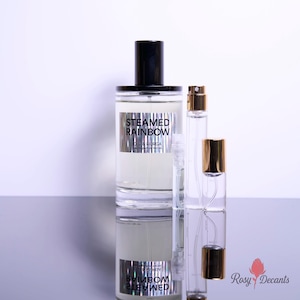May include: A large clear glass perfume bottle with a black cap and a silver label reading STEAMED RAINBOW D.S. & DURGA, accompanied by two smaller clear glass spray vials with gold caps, all reflecting on a smooth surface.
