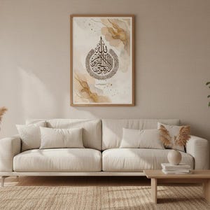 May include: A framed print with Arabic calligraphy in black on a beige and gold abstract background. The artwork is displayed above a beige sofa with matching cushions. A small wooden table and a potted plant are also visible.
