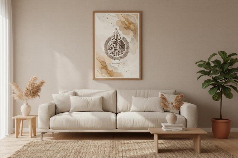 May include: A framed print with Arabic calligraphy in black on a beige and gold abstract background. The artwork is displayed above a beige sofa with matching cushions. A small wooden table and a potted plant are also visible.