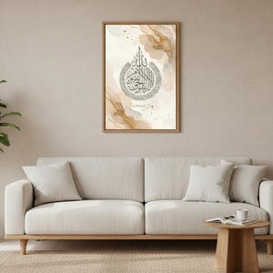 May include: A framed artwork with Arabic calligraphy in a circular design, gold and beige watercolour accents. It hangs above a light-coloured sofa with white cushions. A potted plant and small wooden table complete the room setting.