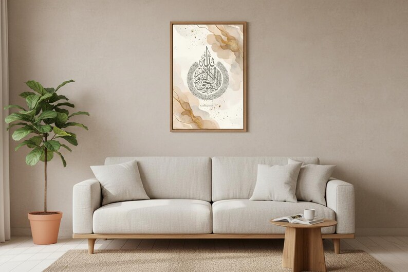 May include: A framed artwork with Arabic calligraphy in a circular design, gold and beige watercolour accents. It hangs above a light-coloured sofa with white cushions. A potted plant and small wooden table complete the room setting.