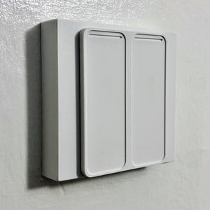 May include: A white light switch with two rectangular buttons. The switch is mounted on a white wall. The design is clean and modern. The switch is likely for controlling lights.