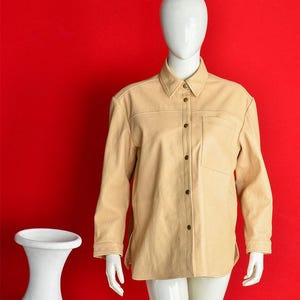 May include: A tan button-down shirt with a collared neckline and a single chest pocket. The long-sleeved shirt is displayed on a mannequin against a red background. The shirt has visible stitching and dark brown buttons.