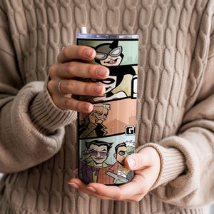 May include: A cylindrical stainless steel tumbler featuring a colorful comic book style graphic print of various characters including a cat-like figure, a jester, and a man with a split face. The tumbler has a clear lid and a white straw.