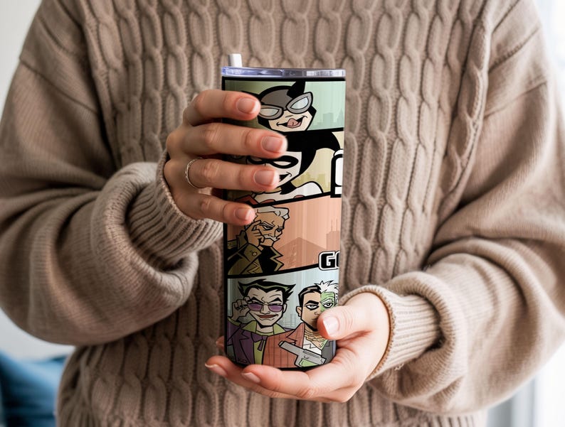 May include: A cylindrical stainless steel tumbler featuring a colorful comic book style graphic print of various characters including a cat-like figure, a jester, and a man with a split face. The tumbler has a clear lid and a white straw.