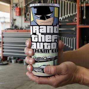 May include: A cylindrical stainless steel tumbler featuring a graphic print of a comic book style character with a mask, the text Grand Theft Auto Gotham City, a black vehicle with fins, and a police car with blue and red lights on the roof.