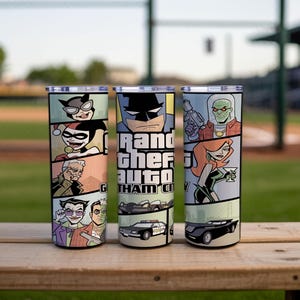 May include: Three cylindrical tumblers with lids featuring a comic book style graphic wrap. The design includes illustrations of various characters, a police car, and the text Grand Theft Auto Gotham City in a bold, blocky font style.