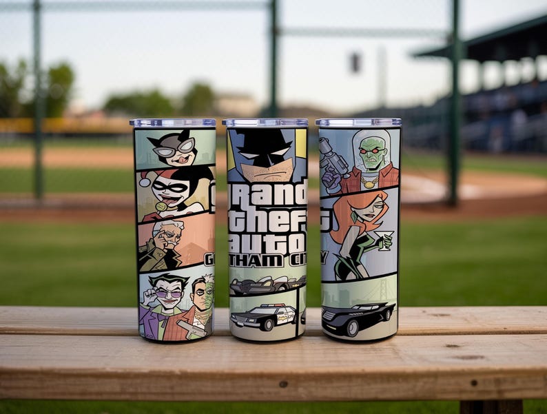 May include: Three cylindrical tumblers with lids featuring a comic book style graphic wrap. The design includes illustrations of various characters, a police car, and the text Grand Theft Auto Gotham City in a bold, blocky font style.