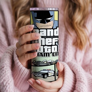 May include: A cylindrical stainless steel tumbler featuring a graphic print of a comic book style Batman character at the top, the text Grand Theft Auto Gotham City in the center, and illustrations of a black sports car and a police car below.
