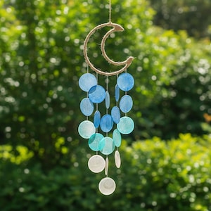 May include: A hanging wind chime featuring a crescent moon shape made of rope. Below the moon are multiple strings of blue, teal, and white circular shell discs. The wind chime is suspended by a thin rope, with a blurred green background.