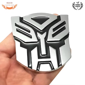 May include: A silver and black emblem with a geometric design, held in a hand. The emblem features a stylized face-like shape with sharp angles and a glossy finish. The design is a recognizable symbol.