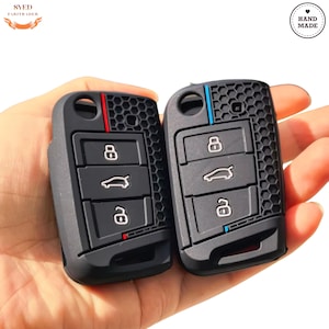 May include: Two black car key fobs with a honeycomb pattern and red and blue accent stripes. Each fob has three buttons with car and lock symbols. The keys are labeled "Hand Made".