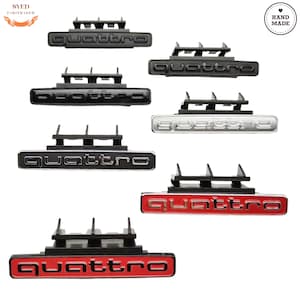 Op de afbeelding: Een verzameling Quattro-auto-emblemen in verschillende kleuren, waaronder zwart, zilver en rood. Elk embleem toont het woord "Quattro" in een vetgedrukte, gestileerde letter, gemonteerd op een zwarte basis met montageclips.