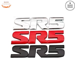 Puede incluir: Tres emblemas SR5 dispuestos verticalmente. El emblema superior es de cromo reflectante, el emblema central es de color rojo brillante sólido y el emblema inferior es de color negro mate. Cada emblema presenta el texto SR5 en negrita.