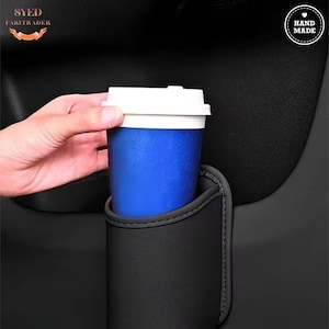 Könnte beinhalten: Ein blauer Einweg-Kaffeebecher mit weißem Deckel in einem schwarzen Getränkehalter im Auto. Der Getränkehalter besteht aus schwarzem, lederähnlichem Material und ist an einem Autositz befestigt. Er hält Getränke sicher.