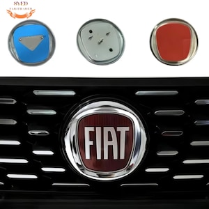 May include: Close-up of a Fiat car emblem and replacement parts. The emblem is a red and silver circular design with the word "FIAT" in white. The replacement parts include a blue emblem, a silver emblem, and a red emblem.