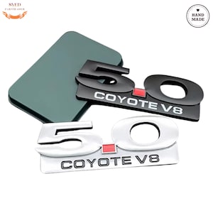Puede incluir: Dos emblemas de coche, uno negro y otro plateado, con el número "5.0", la palabra "COYOTE" y "V8". El emblema negro tiene un acento rojo. El emblema plateado tiene un acento rojo. Los emblemas están sobre una superficie gris.
