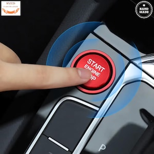 Op de afbeelding: Een rode "START ENGINE STOP" knop wordt ingedrukt. De knop bevindt zich op een zwart en zilveren autoconsole. De afbeelding heeft een blauw cirkelvormig ontwerp rond de knop. De afbeelding heeft ook de tekst "HAND MADE" in een cirkel.