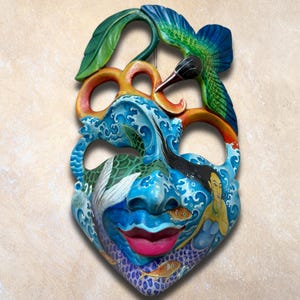May include: Hand-carved wooden mask with a vibrant design. The mask features a blue face with a pink mouth, a mermaid, and fish. A hummingbird and waves are also depicted. The mask is painted with bright colours and has a unique, artistic style.