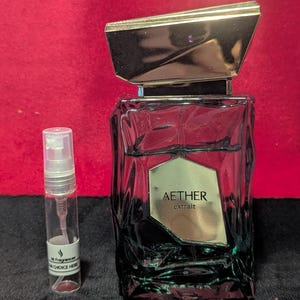 Może przedstawiać: Butelka perfum w kolorze ciemnozielonym ze złotą nakrętką i napisem "AETHER extrait" stoi obok przezroczystej fiolki z próbką. Tło jest czerwone. Butelka perfum ma geometryczny kształt.
