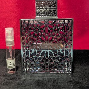 Könnte beinhalten: Eine silberne Parfümflasche mit einem aufwendigen geometrischen Design und dem Wort "RAYHAAN" in Silber. Links ist ein kleines Probenfläschchen. Der Hintergrund ist tiefrot.