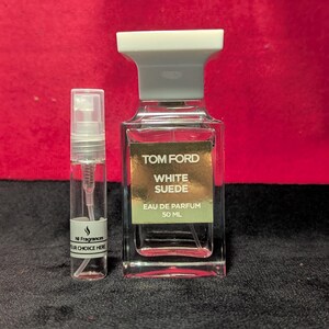 May include: A clear glass perfume bottle with a white cap and the text "TOM FORD White Suede Eau de Parfum 50 ML". A smaller sample spray bottle is next to it. The background is a dark red.