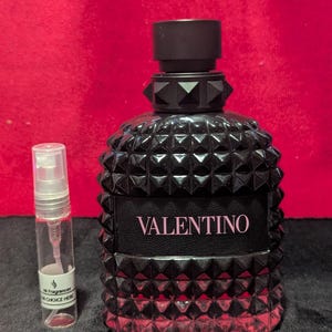 May include: A black perfume bottle with a studded design and the word "VALENTINO" in pink lettering. A small, clear sample vial is next to the bottle. The background is a solid red.