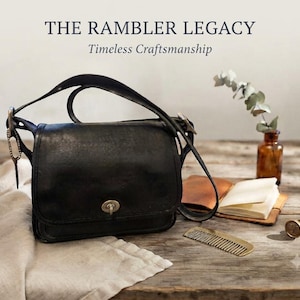 May include: A black leather shoulder bag with a flap closure and a metallic turn-lock mechanism. The text THE RAMBLER LEGACY Timeless Craftsmanship is visible above the bag. The bag features a long strap and is positioned near a small notebook, a comb, and a wax seal stamp.