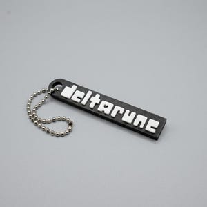 Puede incluir: Un llavero rectangular negro con la palabra "deltarune" en letras blancas. El llavero tiene una cadena de bolas plateada adjunta. El fondo es gris liso.