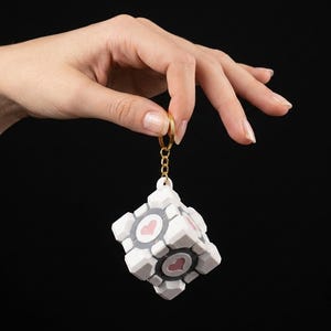 May include: A white cube-shaped keychain featuring a gray circular center with a pink heart design on each face. The cube is attached to a gold-colored metal split ring by a short gold-colored chain, creating a compact and detailed accessory.
