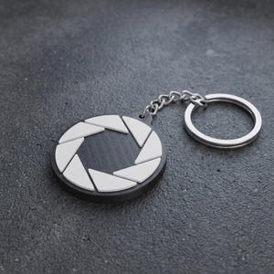 May include: A circular keyring featuring a black base with a white camera aperture design on the front. The object includes a silver-toned metal chain and a split ring for attaching keys, showcasing a clean, geometric aesthetic with high contrast.
