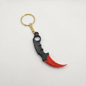May include: A keychain featuring a curved karambit-style blade replica. The handle is matte black with a textured grip and a finger ring at the top, while the blade is a smooth, glossy red. It is attached to a gold-toned metal split ring.