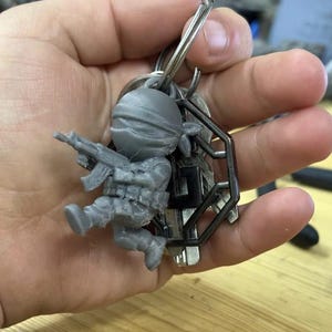 May include: A gray 3D-printed keychain featuring a miniature soldier figure holding a rifle. The soldier is wearing a helmet and tactical gear. The keychain also includes a metal key ring and several keys.