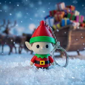 Könnte beinhalten: Ein weihnachtlicher Elfen-Schlüsselanhänger mit roter Mütze, grünem Kragen und rotem Outfit. Der Elf steht in einer verschneiten Szene mit einem Schlitten voller Geschenke im Hintergrund. Rentiere sind ebenfalls in der Ferne zu sehen, was eine festliche Atmosphäre schafft.
