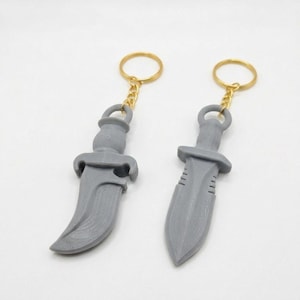 May include: Two gray plastic knife-shaped keychains with gold-colored metal rings. One keychain features a curved blade design, while the other has a straight, double-edged blade. Both items have a textured surface and are attached to gold chains.