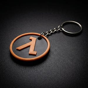 Puede incluir: Un llavero circular que presenta un disco negro con el símbolo griego lambda en relieve de color naranja en el centro. El disco está enmarcado por un borde naranja y está unido a una anilla de metal plateado mediante una cadena corta.
