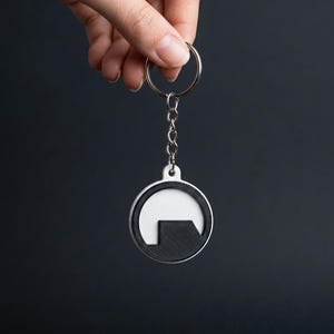 May include: A circular keychain featuring a white outer ring and a black interior design with a stylized lambda symbol. The keychain is attached to a silver-colored metal split ring by a short metal chain, creating a functional accessory.
