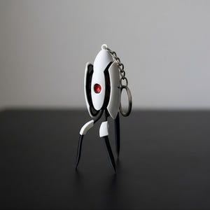 以下が含まれることがあります： 赤色の目を持つ、白と黒のスタイリッシュなロボットデザインのキーホルダー。シルバーのチェーンとリングが付いています。ロボットは3本の黒い脚と丸みを帯びたボディをしています。背景はぼやけたグレーです。