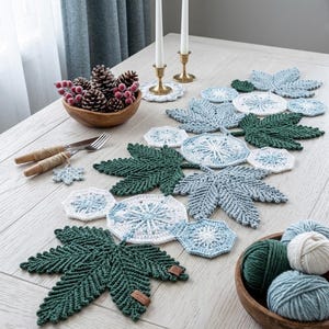 Pode incluir: Uma decoração de mesa com um caminho de mesa em forma de folha de crochê em tons de verde e azul. A mesa é decorada com porta-copos a condizer, uma tigela de madeira com pinhas e castiçais. Há também uma tigela de fio.