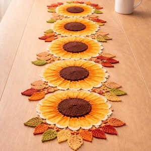 Può includere: Un runner da tavolo all'uncinetto fatto a mano con un motivo a girasole. Il runner presenta quattro grandi girasoli con centri marroni e petali gialli, circondati da foglie all'uncinetto colorate nelle tonalità dell'arancione, del verde e del marrone. Una tazza di caffè bianca è sul tavolo di legno.