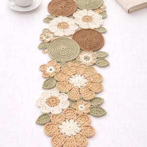Op de afbeelding: Handgemaakte gehaakte tafelloper met een bloemenontwerp in beige, bruin en groen. De loper heeft bloem- en bladmotieven. Een kopje koffie en een boek staan op de achtergrond.