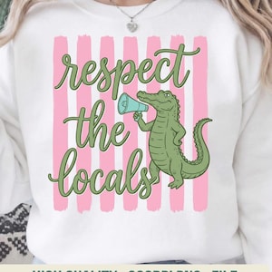 Puede incluir: Sudadera blanca con un diseño gráfico que presenta un caimán verde sosteniendo un megáfono azul. El texto "respect the locals" está escrito en verde sobre rayas verticales rosas.