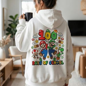 May include: White hoodie featuring a colorful graphic of Mario and Luigi with the text "100 Days of School." The design includes cartoon elements like stars, clouds, and mushrooms. The hoodie is a casual garment.