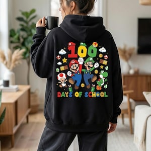 May include: Black hoodie featuring a colorful graphic of Mario and Luigi with the number 100 and the words "DAYS OF SCHOOL". The design includes mushrooms, stars, and other game elements. The hoodie is worn by a person.