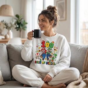 May include: White sweatshirt featuring a colorful graphic of Mario, Luigi, and other characters from the Super Mario Bros. series. The graphic includes the number "100" and the text "Days of School". The sweatshirt is a comfortable, casual style.