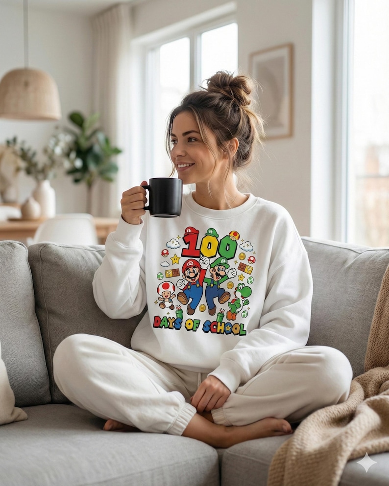 May include: White sweatshirt featuring a colorful graphic of Mario, Luigi, and other characters from the Super Mario Bros. series. The graphic includes the number "100" and the text "Days of School". The sweatshirt is a comfortable, casual style.