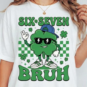 May include: White t-shirt with a green graphic of a four-leaf clover wearing sunglasses and a blue cap. The text "SIX*SEVEN BRUH" is above and below the clover. The design includes green and white checkered squares and shamrocks.
