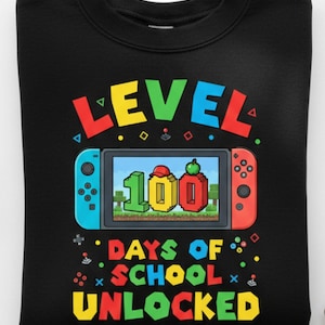 Könnte beinhalten: Schwarzes Sweatshirt mit einer farbenfrohen Grafik, die einen Videospiel-Controller und die Worte "LEVEL 100 DAYS OF SCHOOL UNLOCKED" zeigt. Das Design beinhaltet eine pixelige Spielszene und leuchtende Farben.